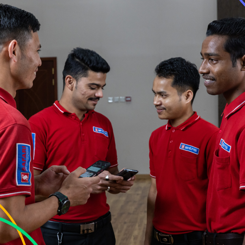 Four workers in red shirts are having a conversation. They are smiling, and two have NOW Money mobile bank app open on their phones.