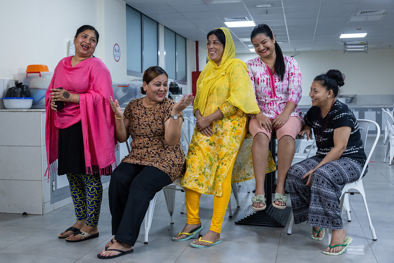 Five women in an office environment, dressed in casual clothing. The women are smiling and laughing.