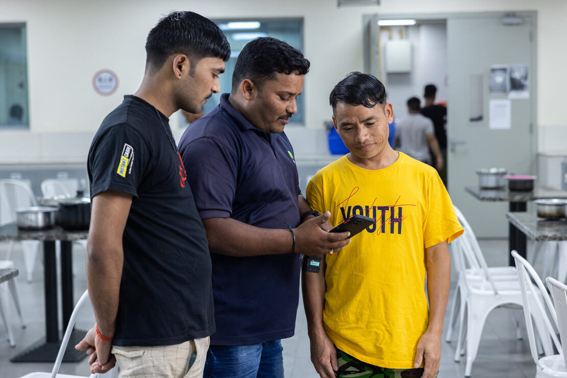 A NOW Employee is demonstrating the NOW Money digital banking app to two migrant workers.